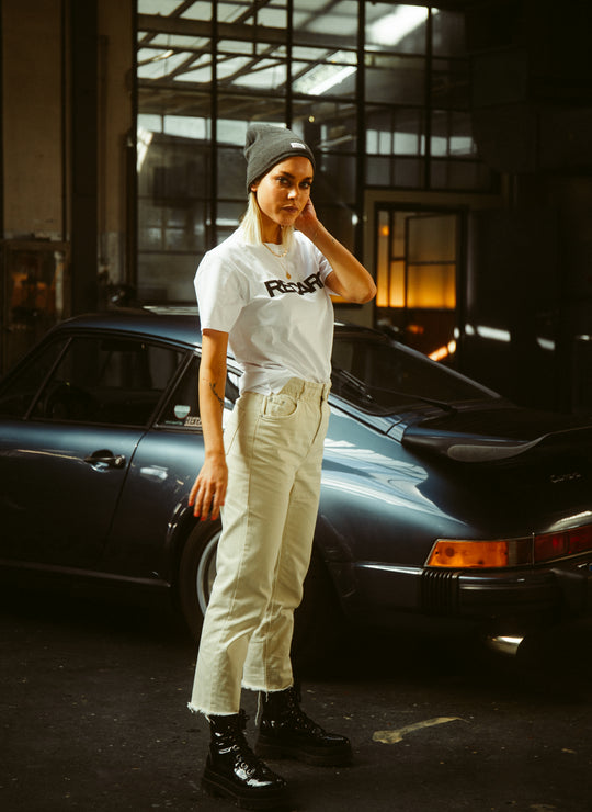 Eine Frau mit grauer Mütze, T-Shirt „Originals RECARO“ von Recaro_Merch_Daniel, cremefarbenen Hosen und schwarzen Stiefeln steht vor einem dunklen Vintage-Sportwagen in einer Garage im Industriestil mit großen Fenstern.