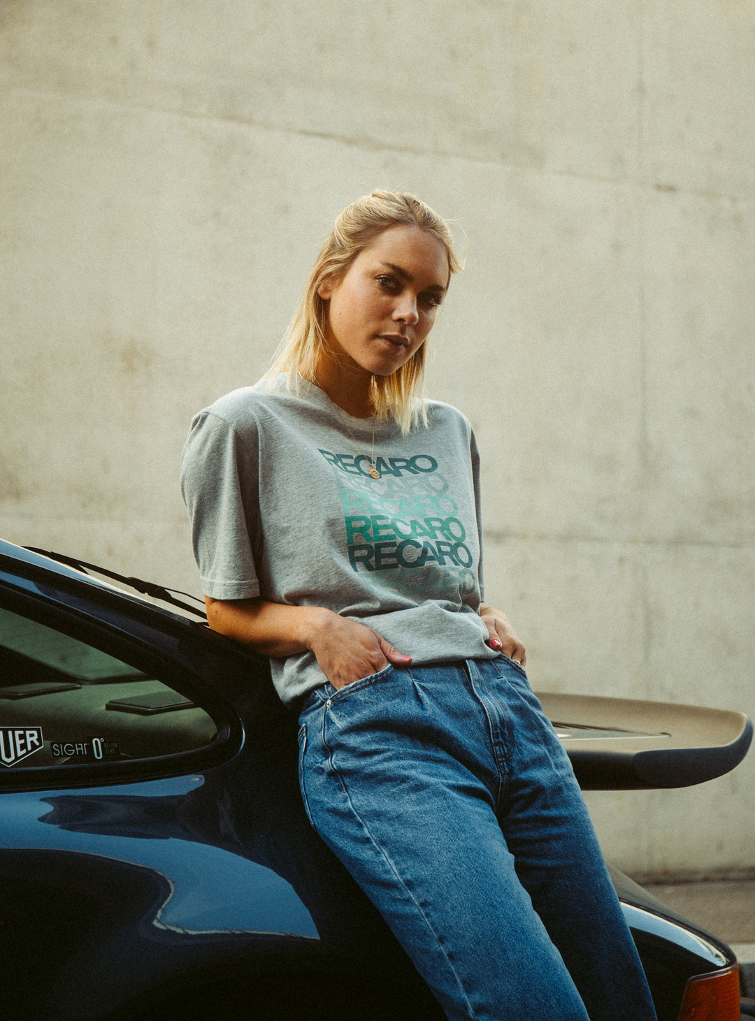 Eine blonde Frau in Bluejeans und einem grauen T-Shirt Spektrum von Recaro lehnt an einem dunkelblauen Auto und präsentiert sich im coolen Retro-Look. Ihr Blick fällt in die Kamera, im Hintergrund ist eine schlichte Betonwand zu sehen.