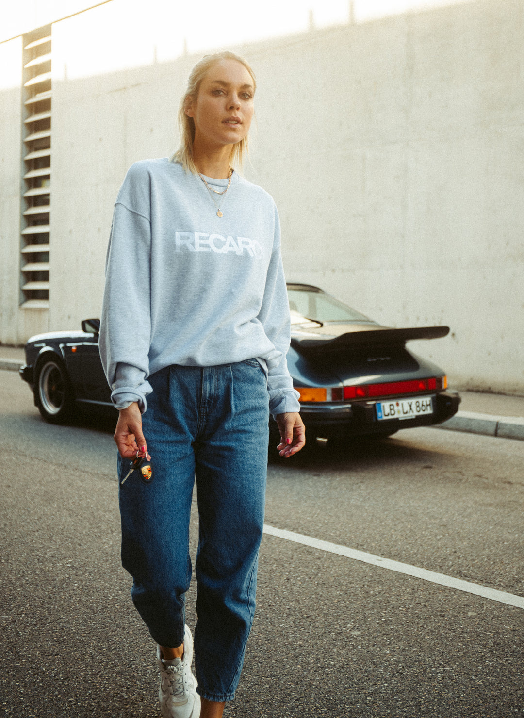 Eine Frau in einem lässigen RECARO Sweatshirt Originals Rubber Logo aus weichem Baumwollmischgewebe und Bluejeans geht mit ihren Autoschlüsseln in der Hand auf einer warm beleuchteten Straße. Hinter ihr parkt ein schwarzer Sportwagen an einer Betonmauer.