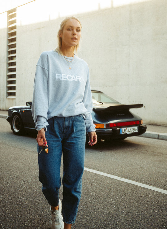 Eine Frau in einem lässigen RECARO Sweatshirt Originals Rubber Logo aus weichem Baumwollmischgewebe und Bluejeans geht mit ihren Autoschlüsseln in der Hand auf einer warm beleuchteten Straße. Hinter ihr parkt ein schwarzer Sportwagen an einer Betonmauer.
