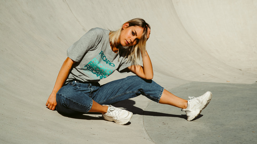 Eine blonde Frau sitzt in einem Skatepark aus Beton auf dem Boden. Sie trägt das Recaro T-Shirt Spektrum, hochgekrempelte Bluejeans für einen Retro-Look und weiße Turnschuhe. Sie berührt ihren Kopf und blickt in die Kamera.