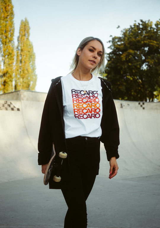 Eine junge Frau mit hellem Haar hält ein Skateboard in der Hand und läuft durch einen Skatepark. Sie trägt ein weißes Recaro T-Shirt Spektrum unter einer schwarzen Jacke. Sie blickt selbstbewusst nach vorne, mit Bäumen und einer Skaterampe im Hintergrund.