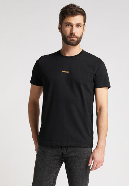 Ein Mann mit kurzen braunen Haaren und Bart trägt das RECARO T-Shirt Backprint mit einem kleinen gelben RECARO-Logo auf schlichter schwarzer Baumwolle, gepaart mit dunklen Jeans, vor einem hellgrauen Hintergrund.