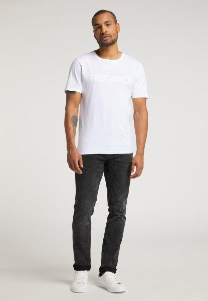 Ein Mann mit kurzen Haaren, gestutztem Bart und einem Tattoo auf dem linken Unterarm trägt das T-Shirt Originals RECARO, gestylt mit schwarzen Jeans und weißen Sneakers vor schlichtem Hintergrund.