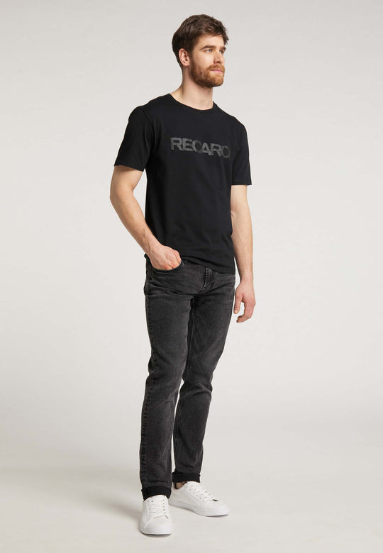 Ein bärtiger Mann steht vor einem schlichten Hintergrund. Er trägt das T-Shirt Originals RECARO von Recaro_Merch_Daniel, dunkle Jeans und weiße Turnschuhe. Eine Hand steckt in der Tasche, der Blick ist leicht zur Seite gerichtet.