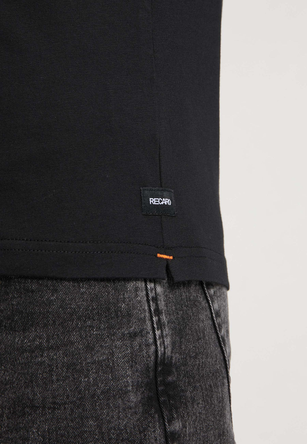 Nahaufnahme einer Person, die das T-Shirt Originals RECARO von Recaro_Merch_Daniel trägt, aus Baumwolle mit orangefarbenen Nähten am unteren Rand und einem kleinen RECARO-Label am Saum, kombiniert mit dunkelgrauen Jeans.