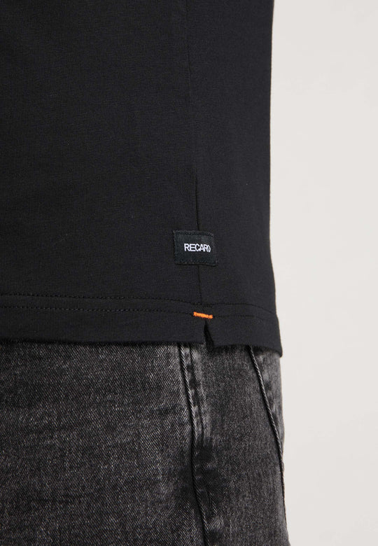 Nahaufnahme einer Person, die das T-Shirt Originals RECARO von Recaro_Merch_Daniel trägt, aus Baumwolle mit orangefarbenen Nähten am unteren Rand und einem kleinen RECARO-Label am Saum, kombiniert mit dunkelgrauen Jeans.
