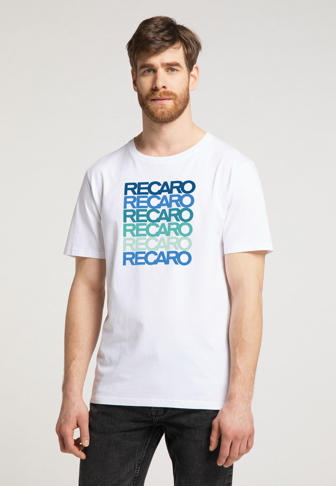 Ein Mann mit braunen Haaren und Bart trägt das weiße „T-Shirt Spektrum“ von Recaro, auf dem fünf RECARO Logos in Blau-Grün-Farbverlauf für einen Retro-Look abgebildet sind. Er steht vor einem schlichten hellen Hintergrund und blickt in die Kamera.