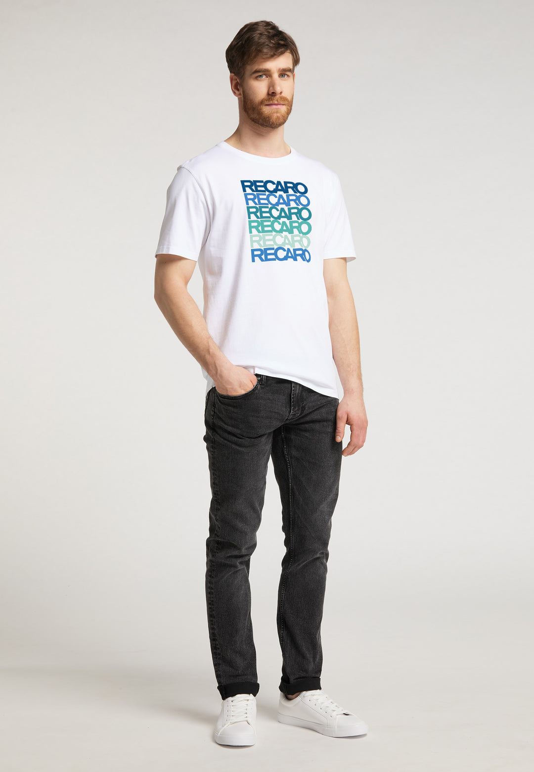 Ein Mann mit Bart und kurzen braunen Haaren posiert in einem weißen Recaro T-Shirt Spektrum, schwarzen Jeans und weißen Sneakers vor hellem Hintergrund, eine Hand in der Tasche für einen coolen Retro-Look.