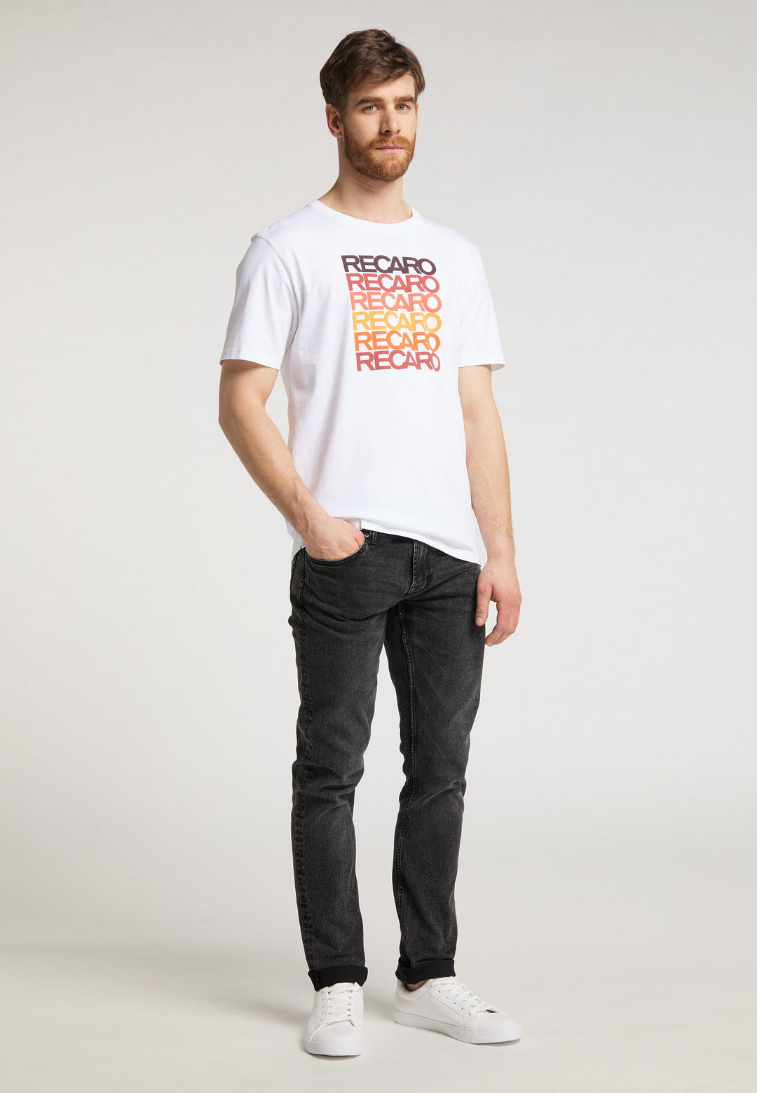 Ein Mann mit hellbraunem Haar und Bart steht vor einem schlichten Hintergrund und trägt das Recaro  T-Shirt Spektrum mit rot-gelbem RECARO-Aufdruck für einen mutigen Retro-Look, kombiniert mit schwarzen Jeans und weißen Turnschuhen.