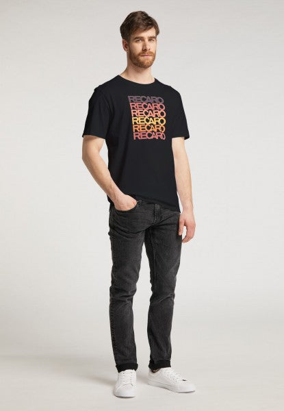 Ein Mann mit kurzen braunen Haaren und Bart präsentiert vor schlichtem hellem Hintergrund das RECARO Spektrum-Bundle – bestehend aus T-Shirt und Snapback-Cap –, gestylt mit schwarzen Jeans und weißen Sneakers für einen coolen Retro-Look.