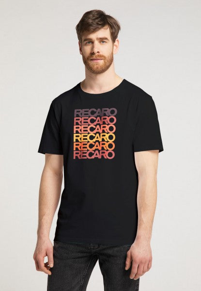 Ein hellhäutiger, braunhaariger Mann mit Bart trägt das RECARO T-Shirt Spektrum in Schwarz, auf dem fünfmal „RECARO“ in Retro-Farbverlauf in Gelb, Orange und Rot abgebildet ist. Er steht vor einem schlichten hellen Hintergrund.