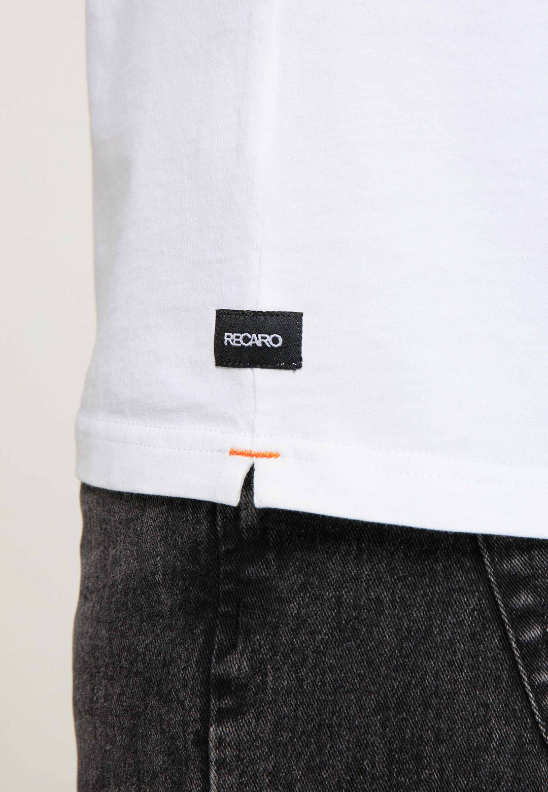 Nahaufnahme eines weißen T-Shirts Spektrum von Recaro mit kleinem schwarzen Etikett am Saum, kombiniert mit dunkler Jeans. Der Fokus liegt auf dem Etikett und dem Saum und verleiht dem Outfit einen dezenten Retro-Look.