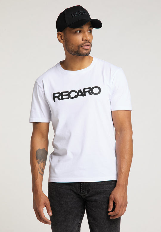 Ein bärtiger Mann mit einem Tattoo auf dem linken Unterarm trägt das RECARO Baseball Cap Originals und ein weißes T-Shirt mit fettem RECARO-Schriftzug, steht vor einem schlichten Hintergrund und blickt zur Seite.