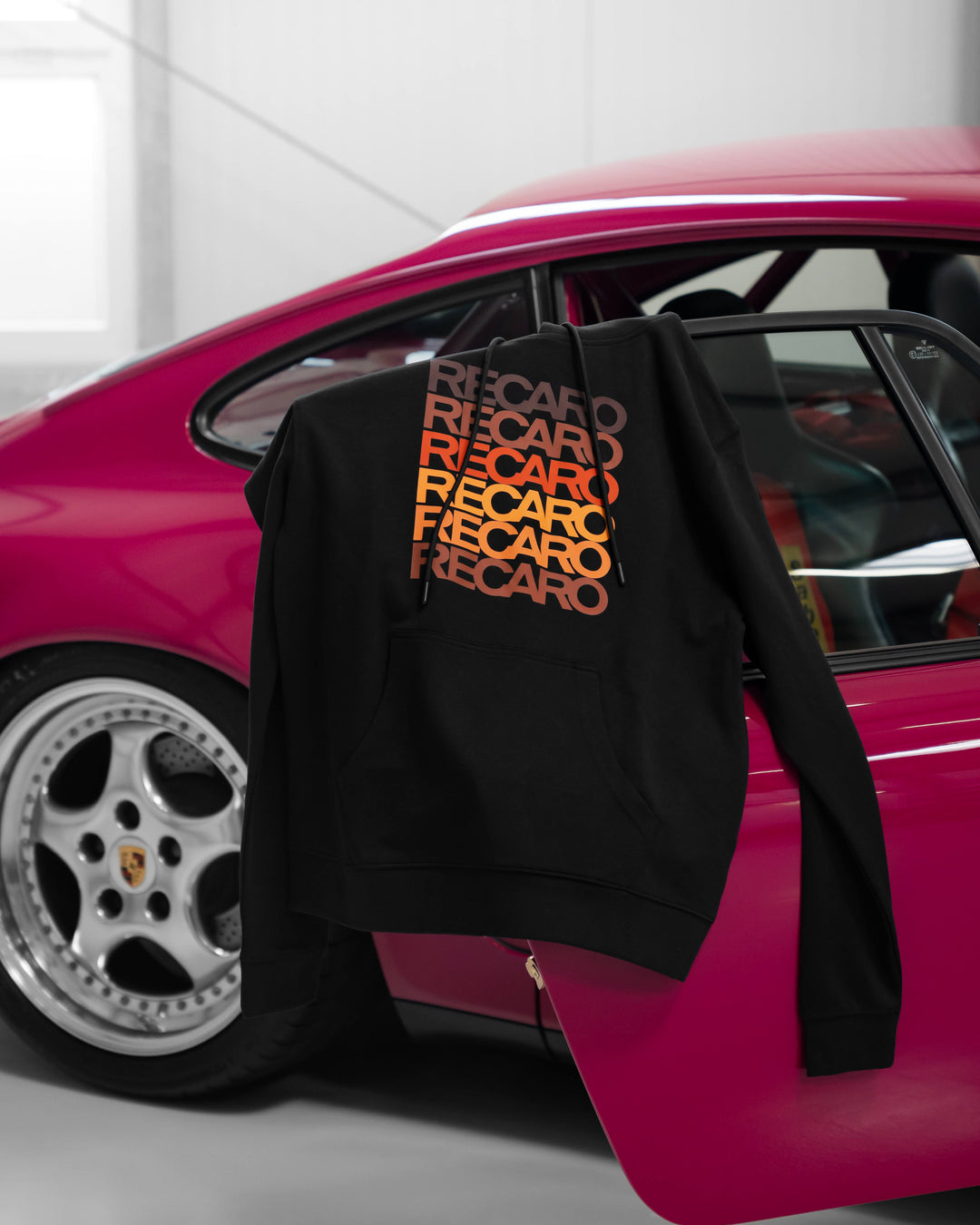Ein schwarzer Kapuzenpullover mit dem RECARO-Aufdruck in Orange und Rot hängt an der offenen Tür eines pinkfarbenen Porsche-Sportwagens, im Hintergrund ist das silberne Lenkrad des Autos zu sehen.