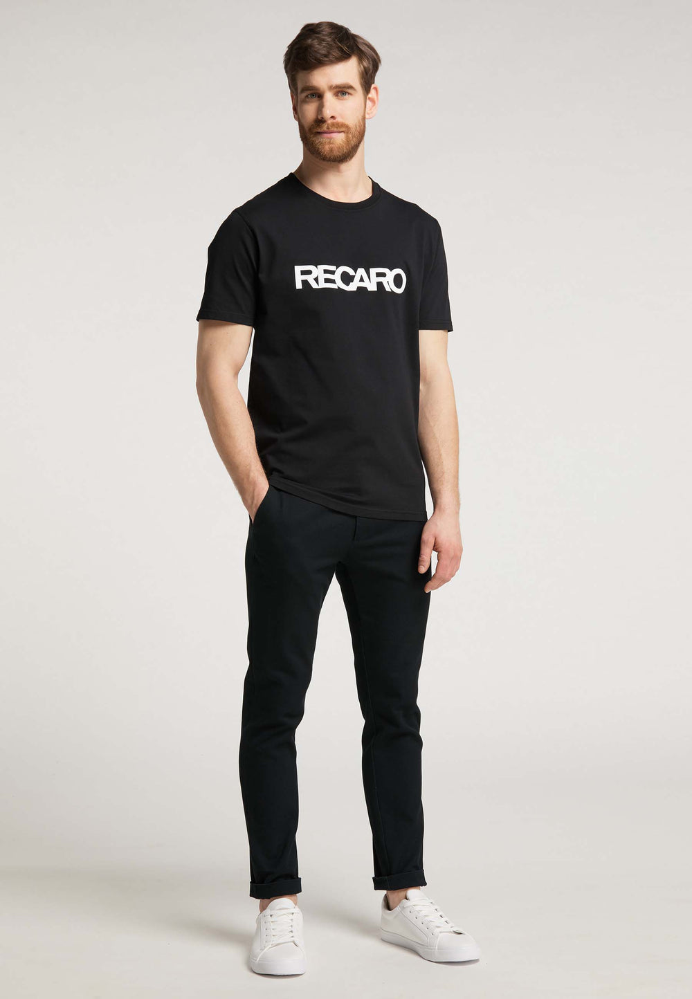 Ein Mann mit kurzen braunen Haaren und Bart trägt ein Recaro T-Shirt Originals RECARO, schwarze Hosen und weiße Turnschuhe und steht mit den Händen in den Taschen vor einem schlichten hellen Hintergrund.