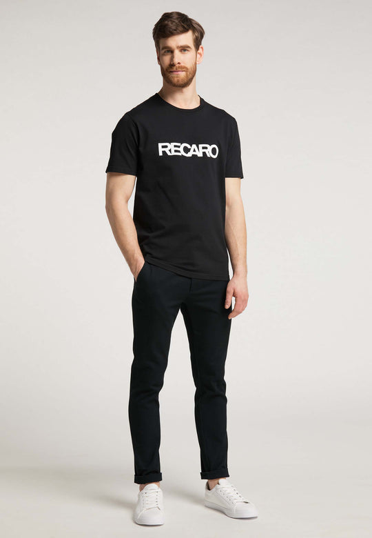Ein Mann mit kurzen braunen Haaren und Bart trägt ein Recaro T-Shirt Originals RECARO, schwarze Hosen und weiße Turnschuhe und steht mit den Händen in den Taschen vor einem schlichten hellen Hintergrund.
