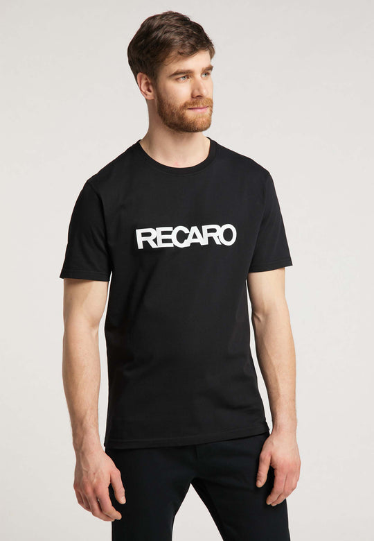 Ein Mann mit braunen Haaren und Bart trägt das T-Shirt Originals RECARO von Recaro. Das auffällige weiße Logo kontrastiert mit dem schwarzen T-Shirt. Er steht vor einem schlichten hellen Hintergrund und blickt leicht zur Seite.