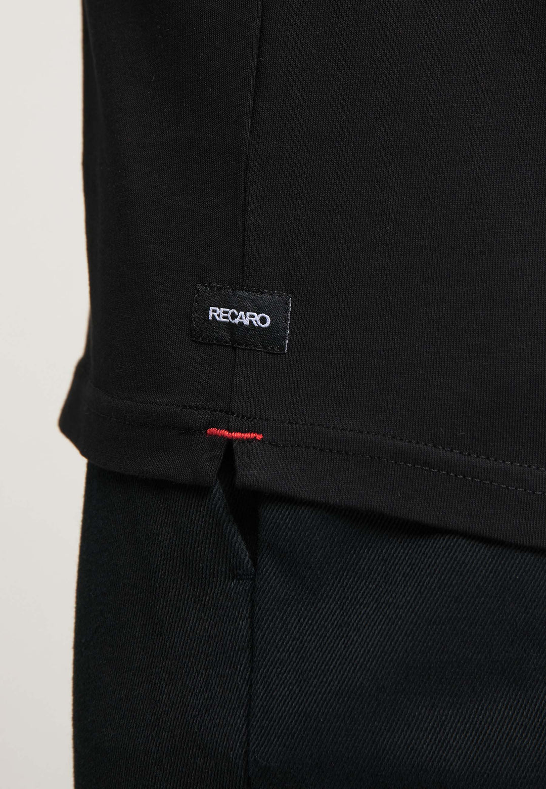 Nahaufnahme eines schwarzen T-Shirts Originals RECARO von Recaro das ein kleines schwarzes RECARO-Etikett mit roten Nähten am Saum und einen Teil der schwarzen Hose darunter zeigt.
