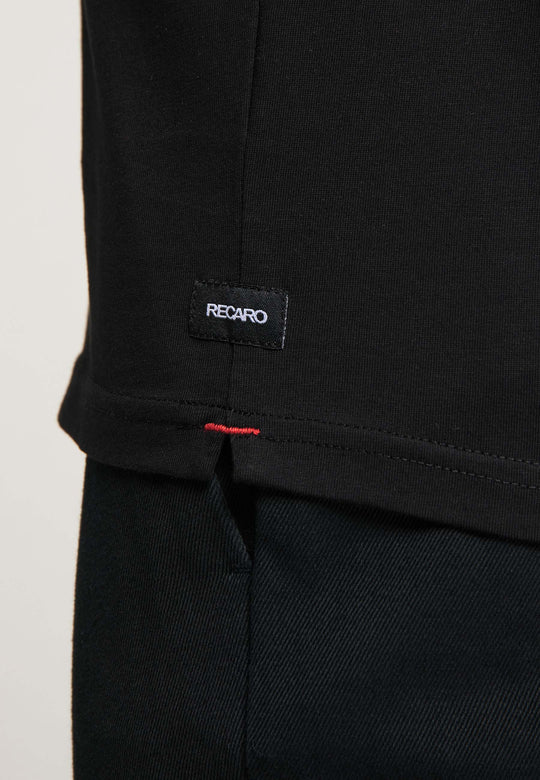 Nahaufnahme eines schwarzen T-Shirts Originals RECARO von Recaro das ein kleines schwarzes RECARO-Etikett mit roten Nähten am Saum und einen Teil der schwarzen Hose darunter zeigt.