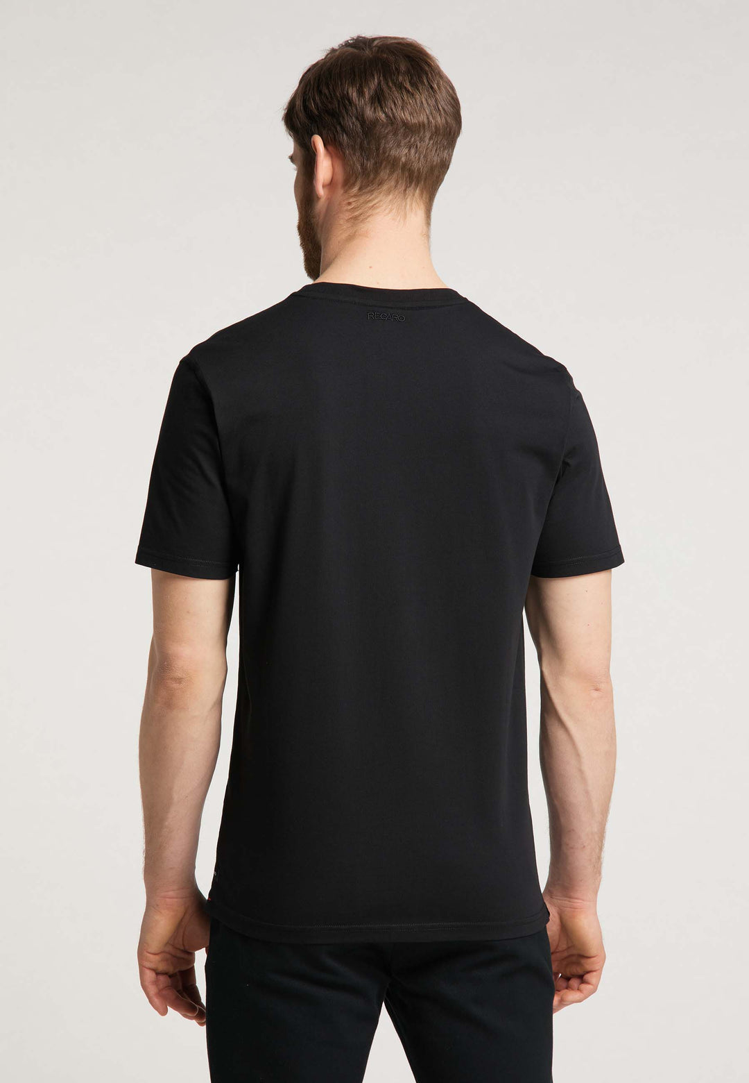 Ein Mann mit kurzen braunen Haaren steht mit dem Rücken zur Kamera und trägt ein T-Shirt Originals RECARO von Recaro und eine dunkle Hose vor einem neutralen Hintergrund.