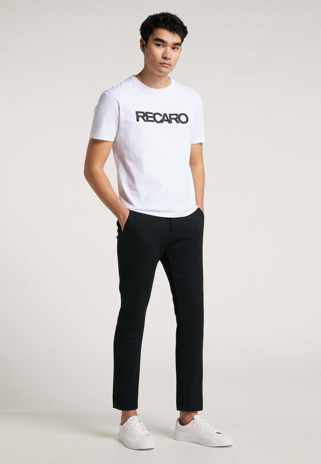 Eine Person steht mit den Händen in den Taschen und trägt ein T-Shirt Originals RECARO von Recaro_Merch_Daniel, schwarze Hosen und weiße Turnschuhe vor einem schlichten hellen Hintergrund.