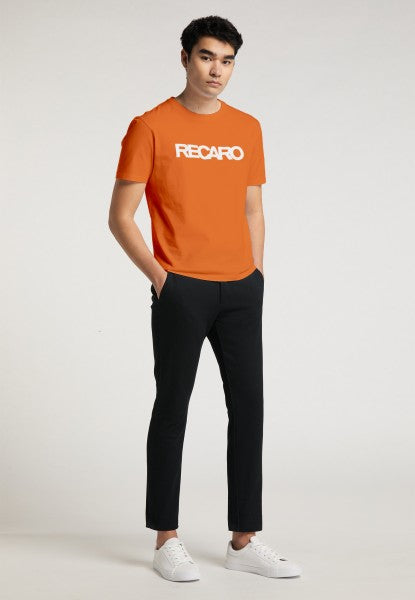 Ein junger Mann steht mit den Händen in den Taschen da und trägt ein orangefarbenes Regular-Fit-T-Shirt „Originals RECARO“ von Recaro_Merch_Daniel, eine schwarze Hose und weiße Turnschuhe vor einem schlichten hellen Hintergrund.