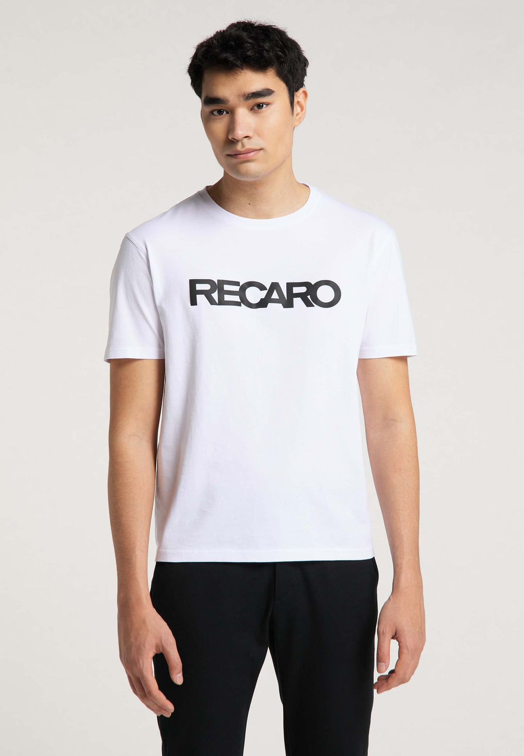 Ein junger Mann mit kurzen dunklen Haaren trägt ein T-Shirt Originals RECARO von Recaro_Merch_Daniel mit einem fetten schwarzen Schriftzug auf der Vorderseite. Er steht mit neutralem Gesichtsausdruck vor einem schlichten, hellen Hintergrund.