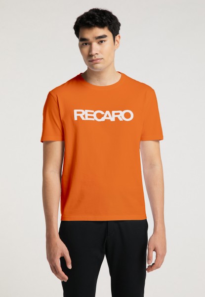 Ein junger Mann mit kurzen dunklen Haaren trägt eine schwarze Hose und ein orangefarbenes Regular-Fit-T-Shirt „Originals RECARO“ von Recaro_Merch_Daniel mit fettem weißen Schriftzug auf der Brust und steht vor einem schlichten Hintergrund.