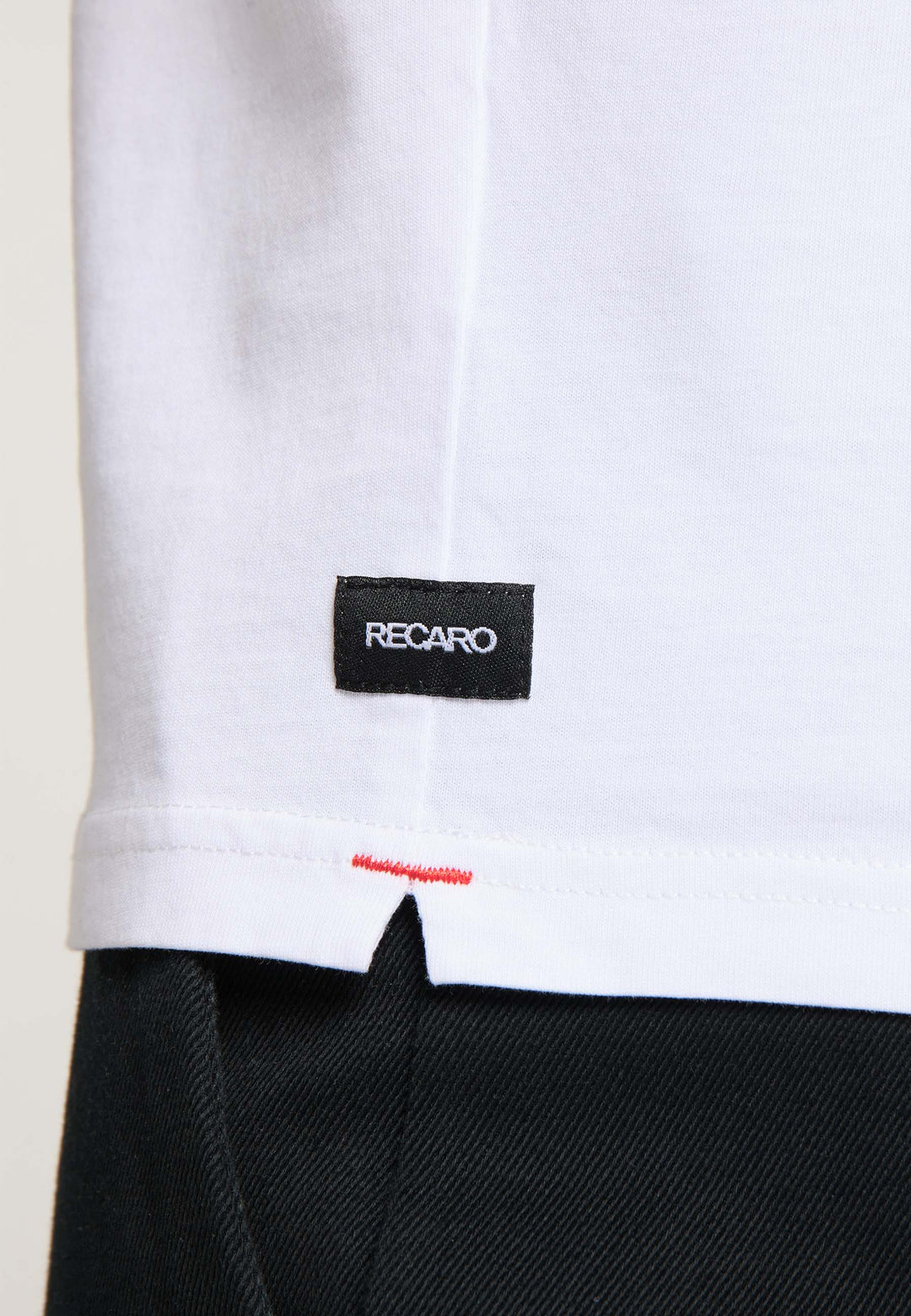 Nahaufnahme des unteren Teils eines T-Shirts Originals RECARO von Recaro_Merch_Daniel aus hochwertiger Baumwolle mit einem kleinen schwarzen „RECARO“-Etikett und roten Nähten über der schwarzen Hose.
