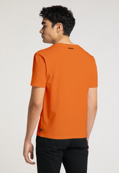 Eine Person mit kurzen dunklen Haaren steht mit abgewandtem Gesicht da und trägt ein orangefarbenes T-Shirt mit normaler Passform „Originals RECARO“ von Recaro_Merch_Daniel und eine schwarze Hose vor einem schlichten hellen Hintergrund.
