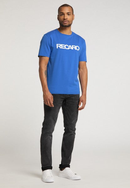 Ein Mann blickt nach vorne und trägt ein leuchtend blaues T-Shirt „Originals RECARO“ von Recaro_Merch_Daniel, kombiniert mit schwarzen Jeans und weißen Turnschuhen vor einem schlichten hellen Hintergrund.