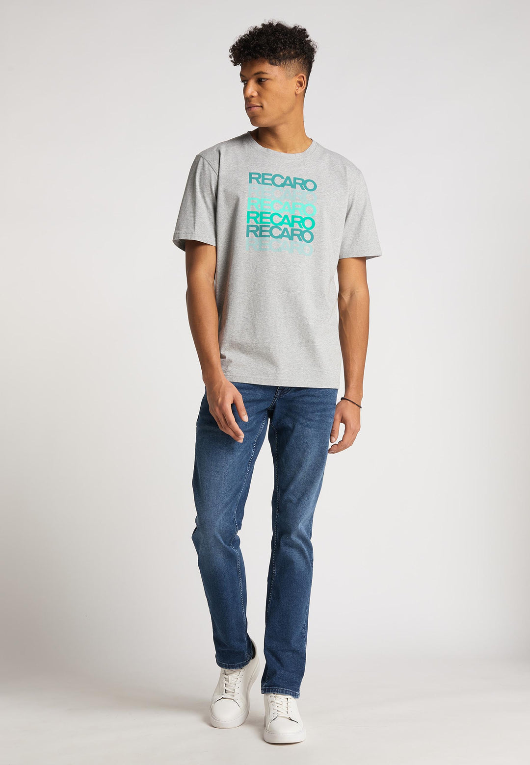 Ein junger Mann steht vor einem schlichten Hintergrund und trägt das graue T-Shirt Spektrum von Recaro aus Baumwolle, blaue Jeans und weiße Turnschuhe. Er blickt zur Seite, eine Hand ruht auf dem Boden, im dezenten Retro-Look.