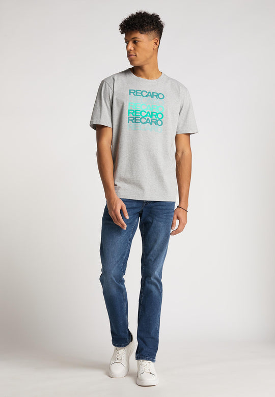 Ein junger Mann steht vor einem schlichten Hintergrund und trägt das graue T-Shirt Spektrum von Recaro aus Baumwolle, blaue Jeans und weiße Turnschuhe. Er blickt zur Seite, eine Hand ruht auf dem Boden, im dezenten Retro-Look.