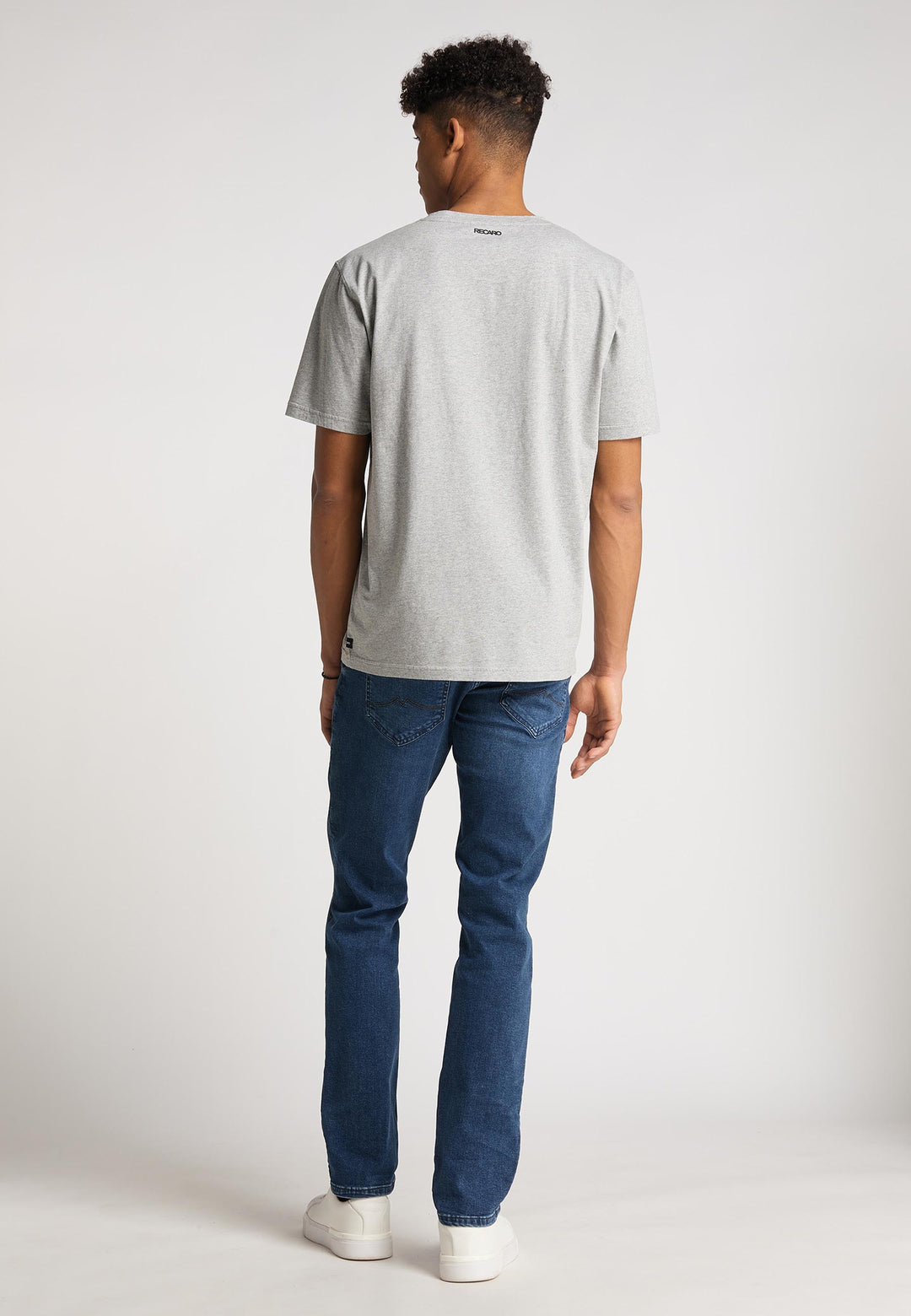 Ein Mann blickt von der Kamera weg und trägt das hellgraue Recaro  T-Shirt Spektrum, Bluejeans und weiße Sneakers im Retro-Look vor einem schlichten hellen Hintergrund.