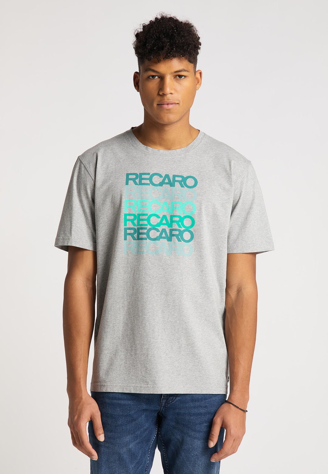 Ein junger Mann trägt das Recaro T-Shirt Spektrum in Hellgrau mit dem sich wiederholenden RECARO-Schriftzug in Blau und Türkis. Mit seinen kurzen, lockigen Haaren und Blue Jeans fällt er vor dem schlichten weißen Hintergrund auf.