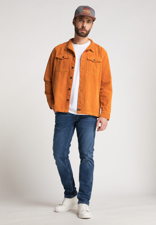 Ein Mann, der die RECARO_Merchandising Snapback Cap Spektrum, eine orangefarbene Jacke mit Knöpfen über einem weißen T-Shirt, blaue Jeans und weiße Turnschuhe trägt, steht vor einem schlichten hellen Hintergrund und blickt mit einem subtilen Retro-Vibe in die Kamera.
