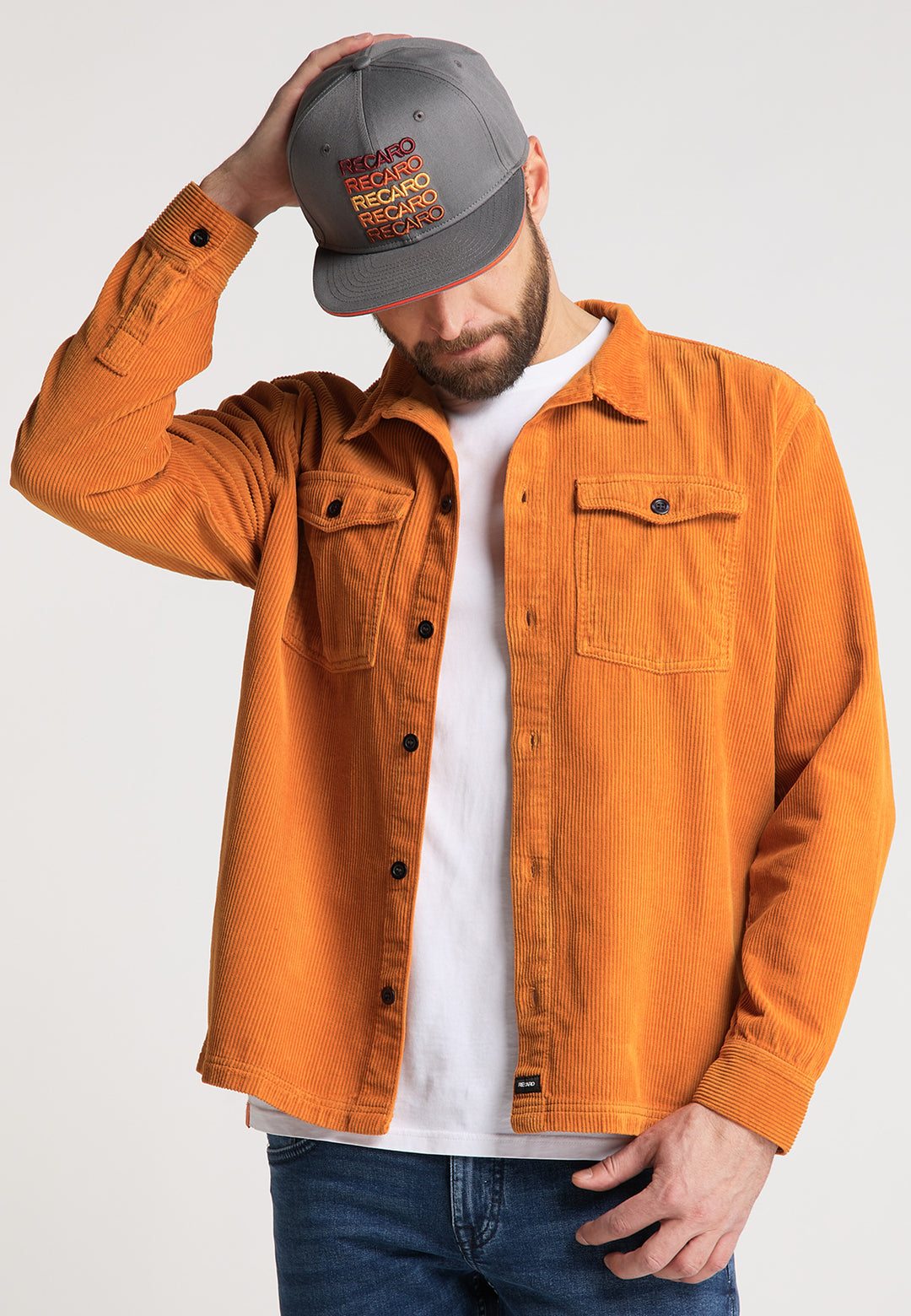 Ein Mann in einer grauen RECARO_Merchandising Snapback Cap Spektrum, einem orangefarbenen Cordhemd mit Knöpfen, einem weißen T-Shirt und Bluejeans steht mit Blick nach vorne da und berührt die Krempe seiner Kappe vor einem schlichten hellen Hintergrund für einen klassischen Retro-Look.