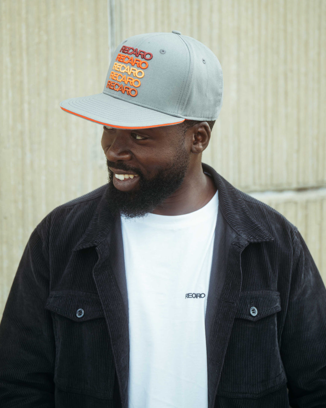 Ein Mann mit Bart trägt eine graue Snapback Cap Spektrum und ein weißes RECARO_Merchandising T-Shirt, kombiniert mit einer dunklen Cordjacke. Er steht vor einer strukturierten beigen Wand und blickt mit einem leichten Lächeln nach links, um ein Retro-Feeling zu erzeugen.