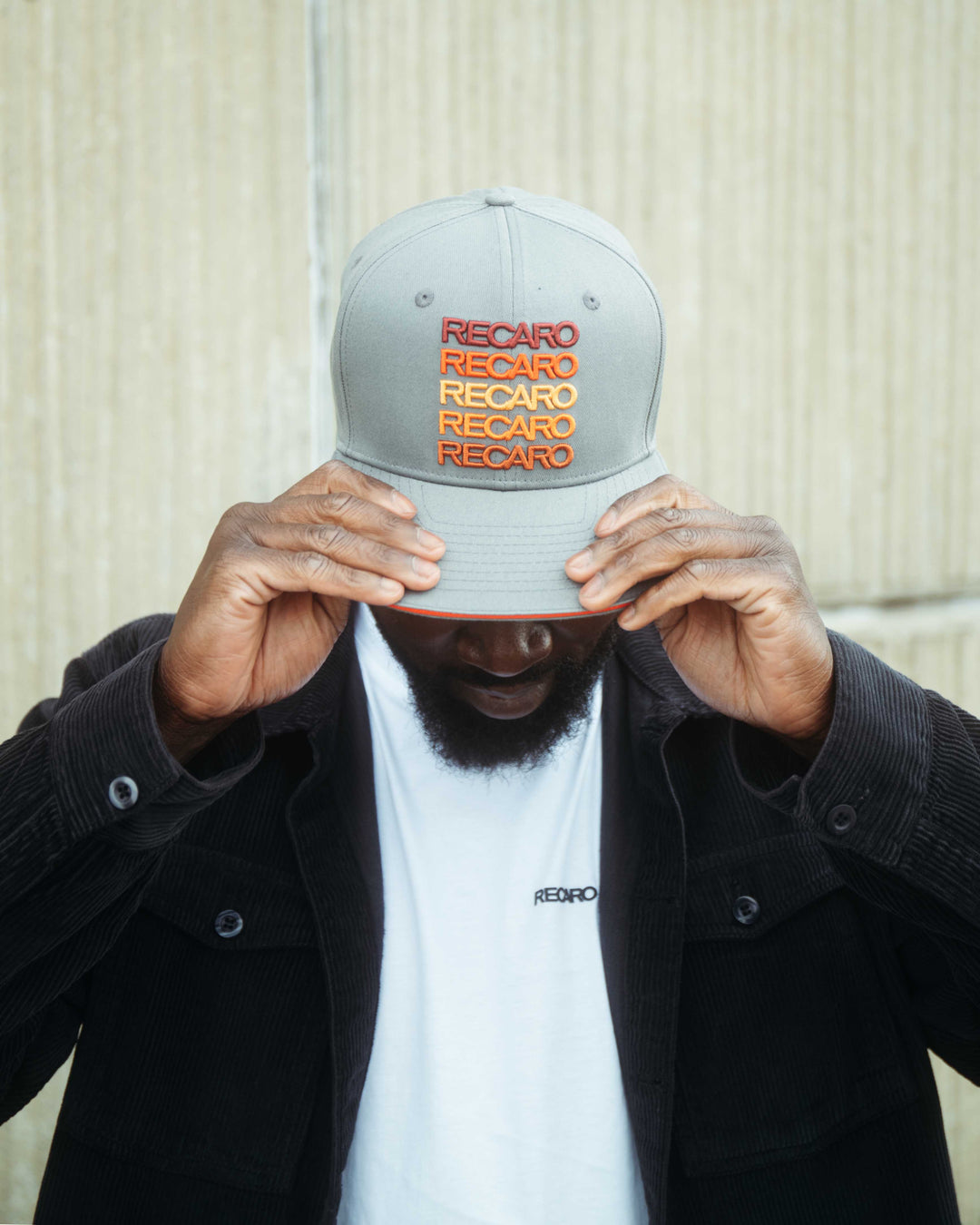 Ein Mann im Retro-Stil trägt eine graue RECARO_Merchandising Snapback Cap Spektrum mit dem Markennamen in Rot und Orange. Er zieht die Krempe tief über die Augen, trägt eine schwarze Jacke und ein weißes RECARO Shirt.