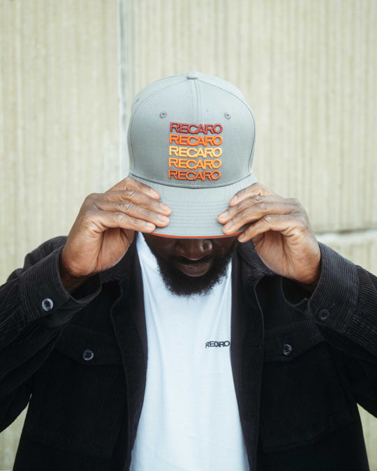Ein Mann im Retro-Stil trägt eine graue RECARO_Merchandising Snapback Cap Spektrum mit dem Markennamen in Rot und Orange. Er zieht die Krempe tief über die Augen, trägt eine schwarze Jacke und ein weißes RECARO Shirt.