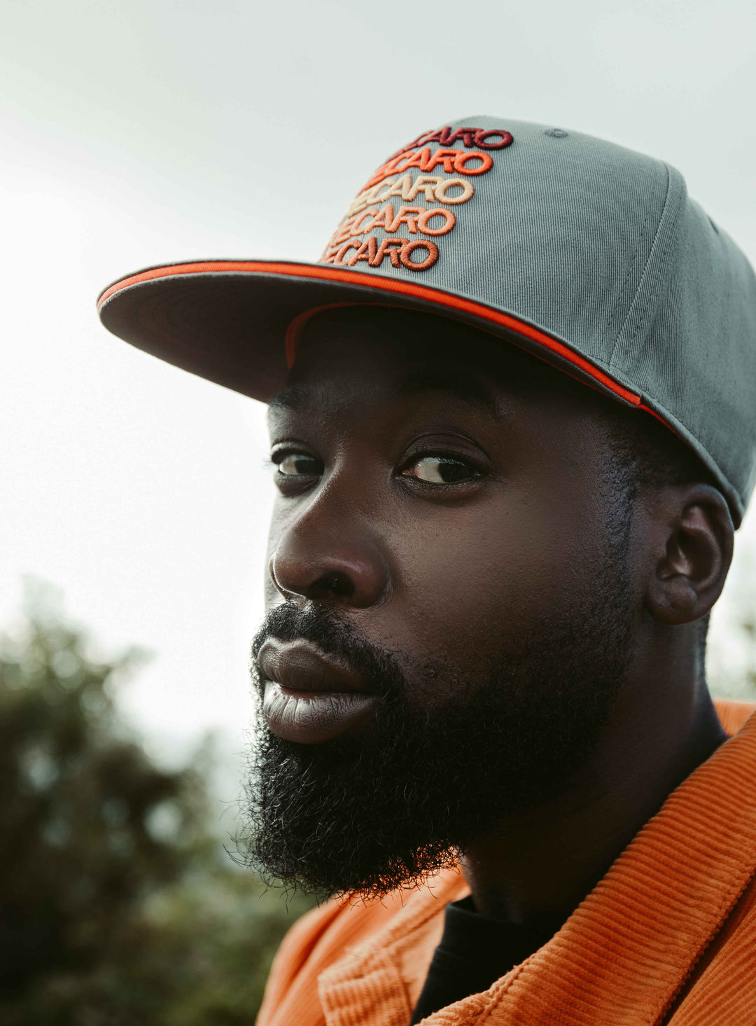 Ein bärtiger Mann mit einem grau-orangen RECARO_Merchandising Snapback Cap Spektrum und einer orangefarbenen Jacke blickt ernst in die Kamera. Der unscharfe Außenhintergrund sorgt für ein dezentes Retro-Feeling.