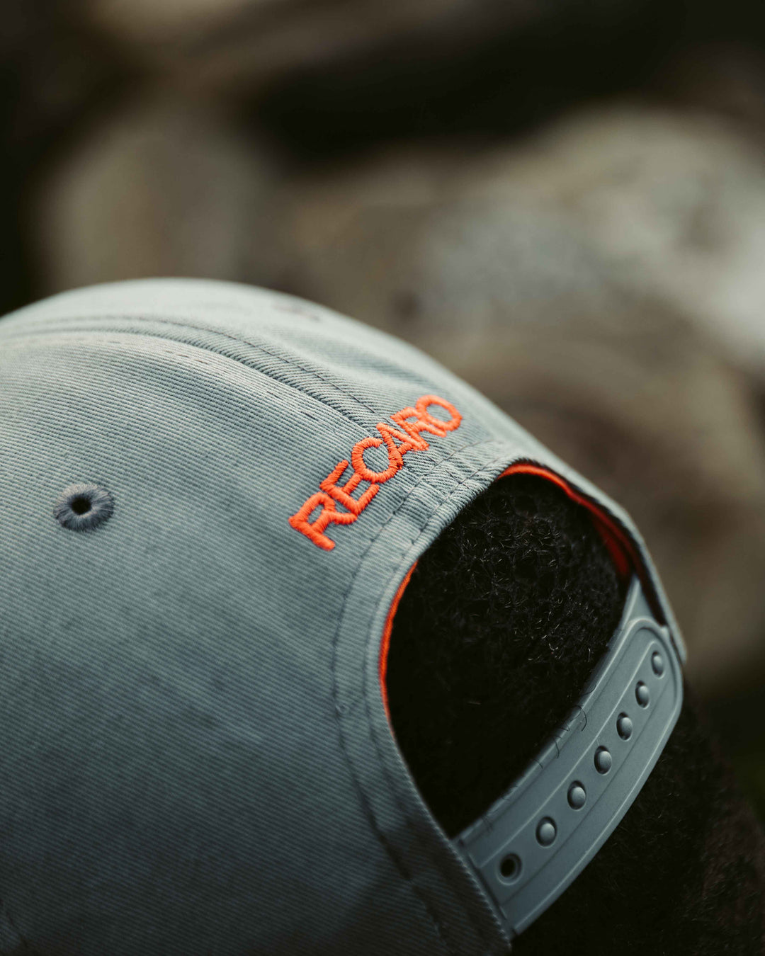 Nahaufnahme der Rückseite einer grauen Snapback-Kappe Spektrum von RECARO_Merchandising mit orangefarbener Stickerei über einem verstellbaren Riemen, getragen von einer Person mit lockigem schwarzen Haar für einen coolen Retro-Look.