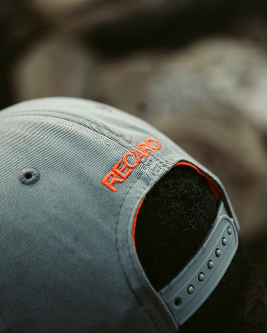 Nahaufnahme der Rückseite einer grauen Snapback-Kappe Spektrum von RECARO_Merchandising mit orangefarbener Stickerei über einem verstellbaren Riemen, getragen von einer Person mit lockigem schwarzen Haar für einen coolen Retro-Look.