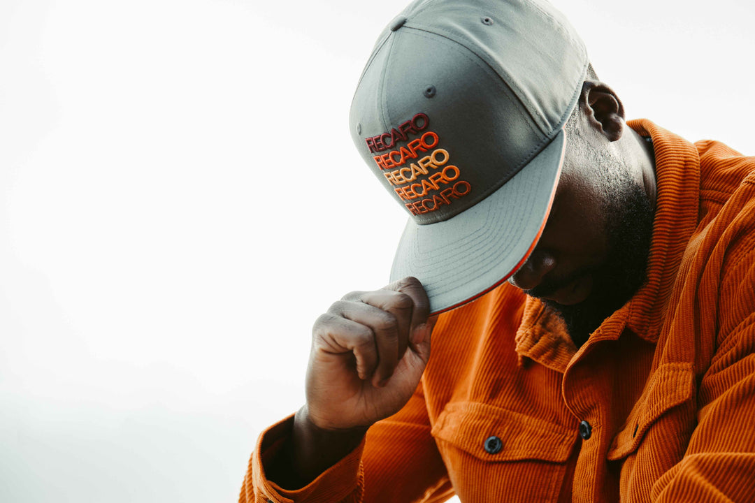 Eine Person in einer orangefarbenen Cordjacke und einem grauen RECARO_Merchandising Snapback Cap Spektrum senkt den Kopf und hält die Krempe fest, wodurch vor einem hellen Hintergrund ein Retro-Look entsteht.