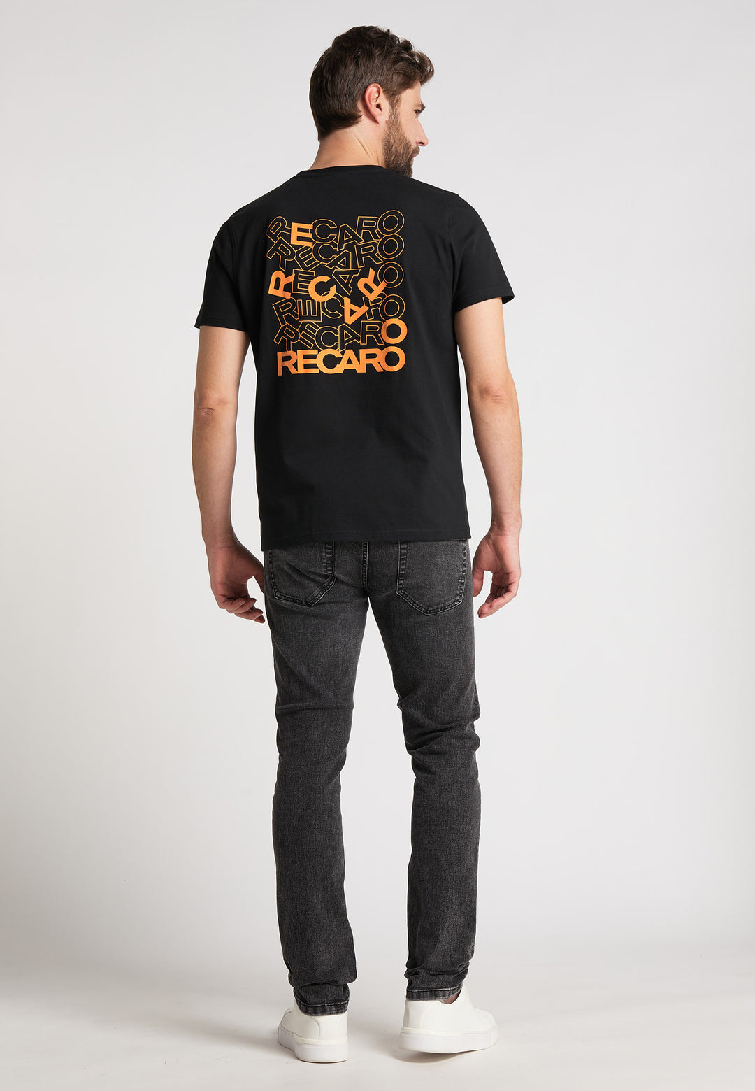 Ein Mann blickt abgewandt in dem RECARO T-Shirt Backprint, einem schwarzen Baumwoll-T-Shirt mit orangefarbenen und gelben RECARO-Logos auf der Rückseite, kombiniert mit dunklen Jeans und weißen Turnschuhen vor einem schlichten hellen Hintergrund.