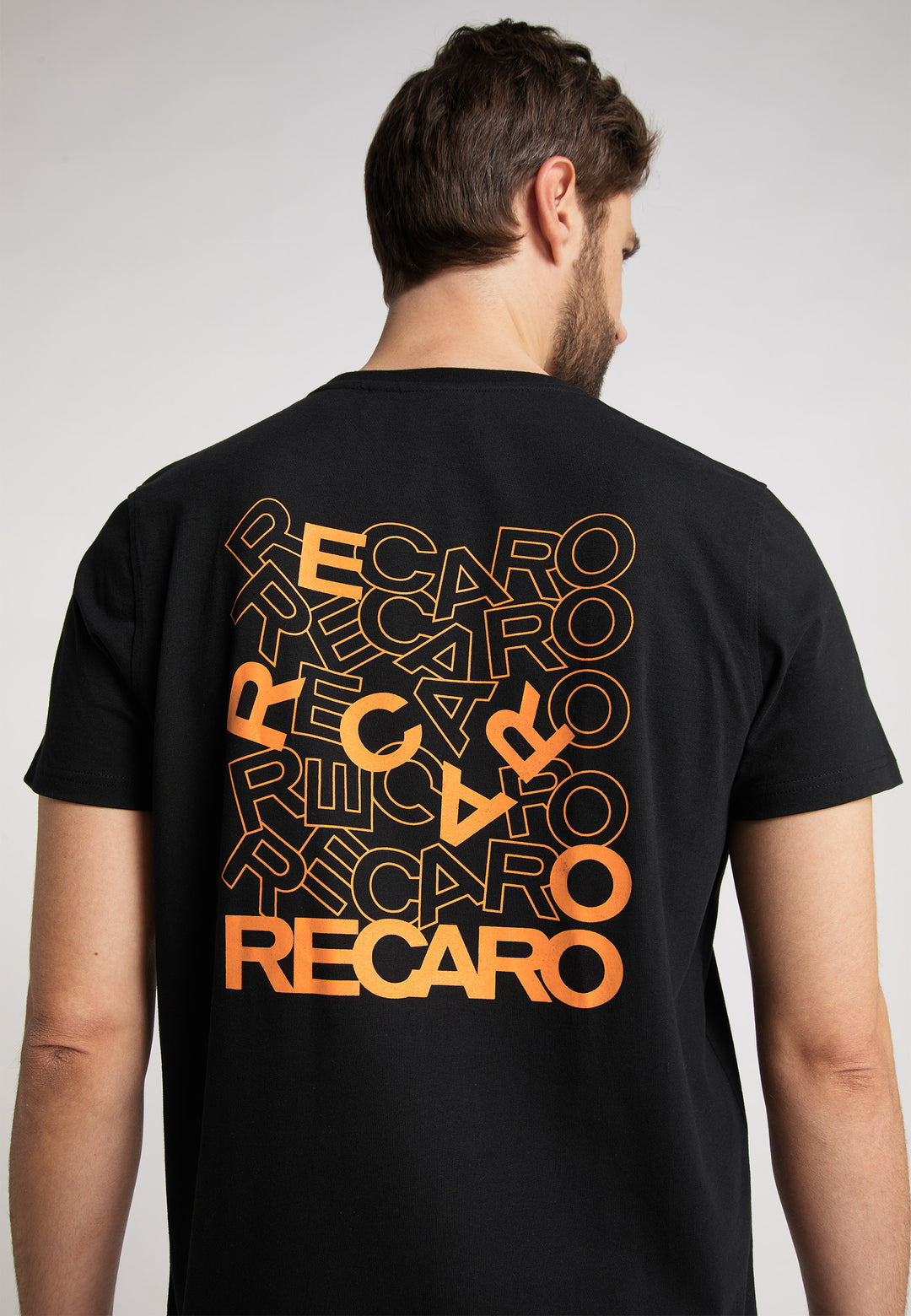 Ein Mann trägt mit dem Rücken zur Kamera das Recaro_Merch_Daniel T-Shirt Backprint – ein schwarzes Baumwoll-T-Shirt mit mehreren orange-gelben RECARO-Logos im Farbverlauf auf der Rückseite.