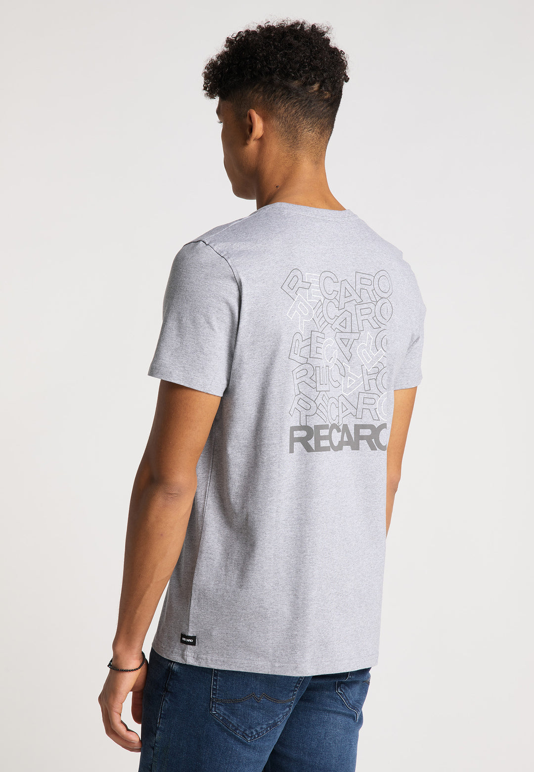 Eine Person in Blue Jeans und mit kurzen lockigen Haaren blickt abgewandt und trägt das RECARO T-Shirt Backprint in Hellgrau aus weichem Baumwollstoff, das die überlappenden Logos auf der Rückseite zur Schau stellt.