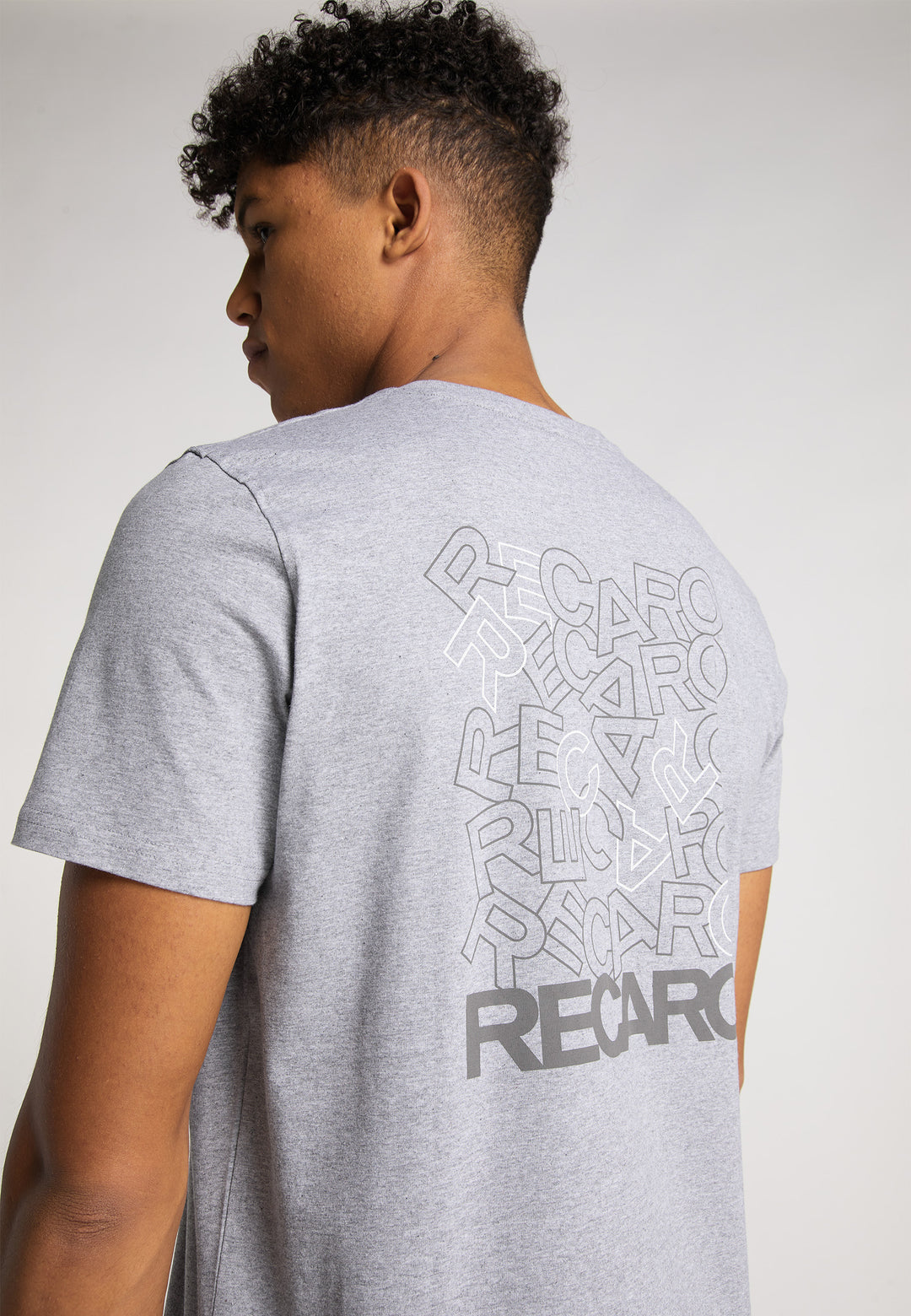 Jemand trägt das RECARO T-Shirt Backprint in Hellgrau, auf dessen Rückseite das RECARO Logo mehrfach in unterschiedlichen Größen und Winkeln aufgedruckt ist, vor einem schlichten hellen Hintergrund.