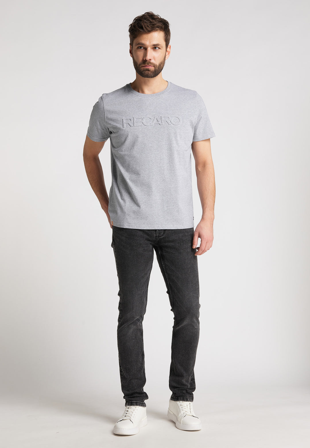 Ein Mann mit kurzen braunen Haaren und Bart steht vor einem schlichten weißen Hintergrund und trägt das hellgraue RECARO T-Shirt Originals Embossed mit einem auffälligen 3D-RECARO-Logo, schwarze Jeans und weiße Turnschuhe.